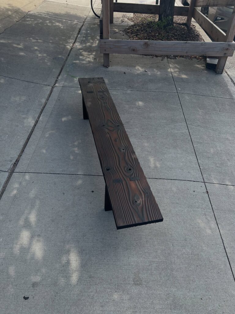 Charred black wooden bench with visible grain on a grey concrete sidewalk.