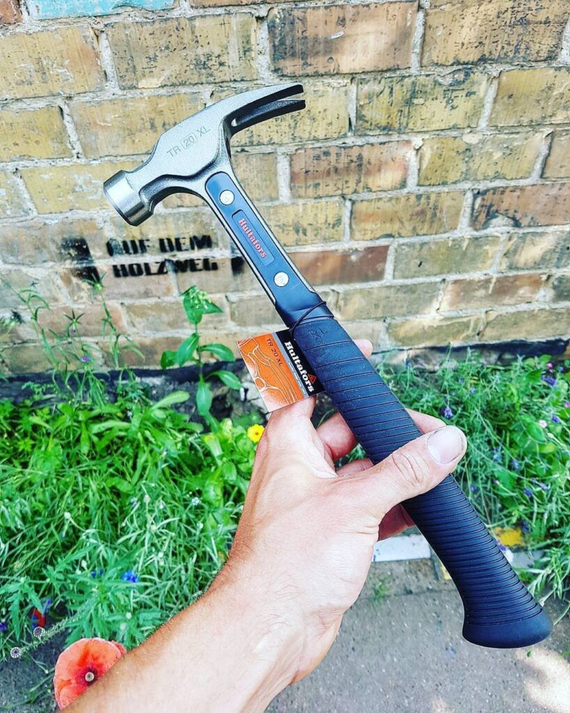 Claw hammer with black grip held by a hand against a brick wall.