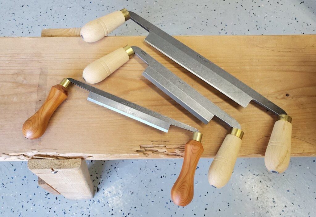 Three drawknives with wooden handles resting on a thick light-colored wooden beam.