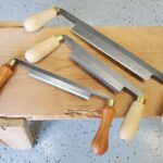 Three drawknives with wooden handles resting on a thick light-colored wooden beam.