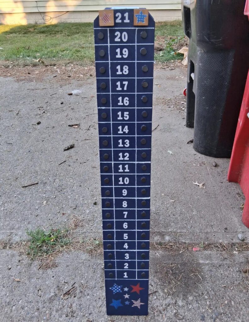 Wooden cornhole scoreboard painted blue with white numbers and pegs on concrete.