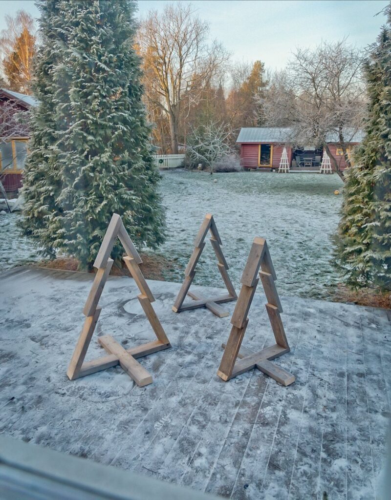 Three minimalist wooden Christmas tree structures on a frost-covered patio in a winter landscape.