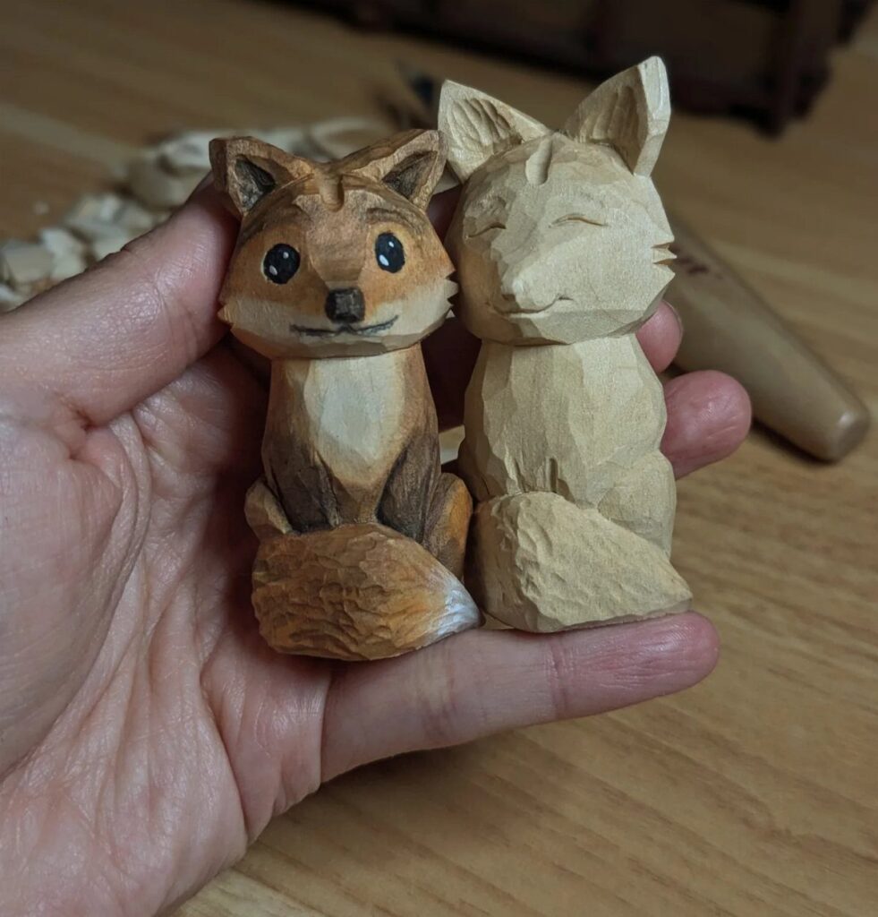 Two small hand-carved wooden foxes, one painted orange and one natural wood, held in hand.