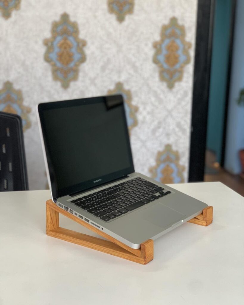 Silver laptop resting on a light-brown wooden ergonomic stand on a white table.